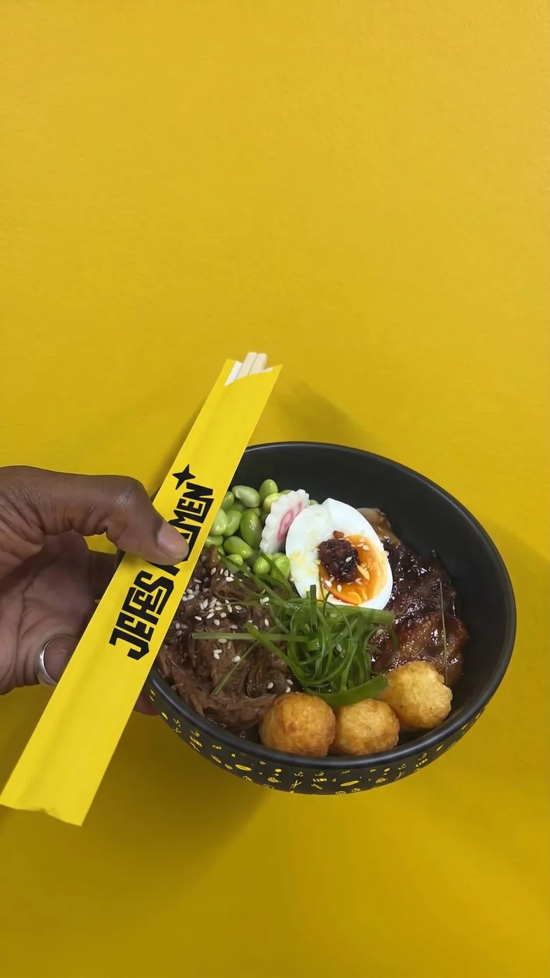 A hand holding a black bowl of ramen topped with pulled beef, a soft-boiled egg with chilli oil, edamame beans, tater tots and spring onion, with a yellow Jefes Ramen chopstick sleeve, against a yellow wall