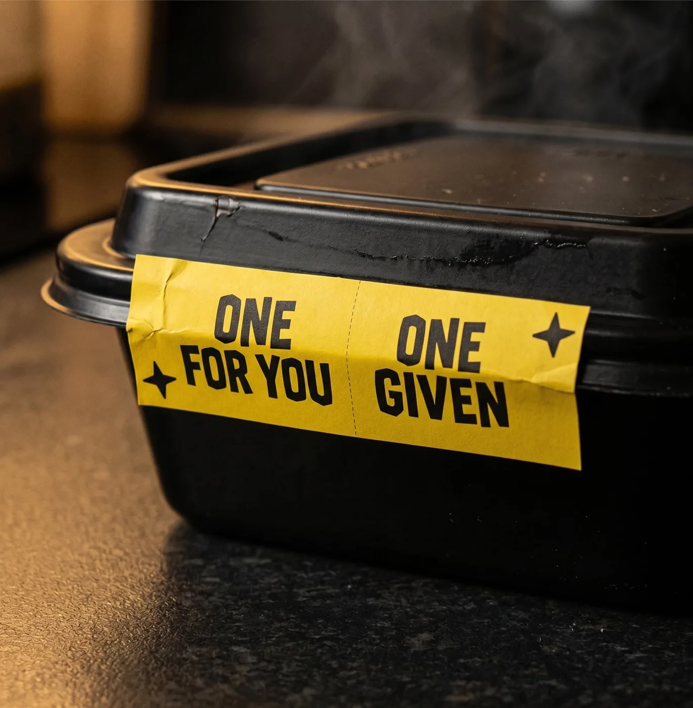 A black takeaway food container on a dark counter with a yellow label reading One for you, One given