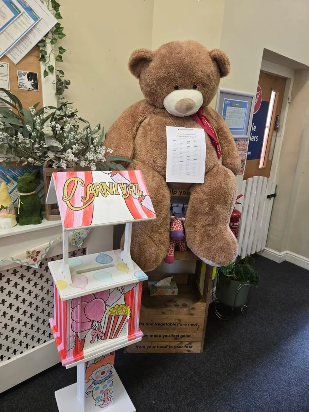 A large brown teddy bear with a note pinned to it, beside a hand-painted wishing well donation box