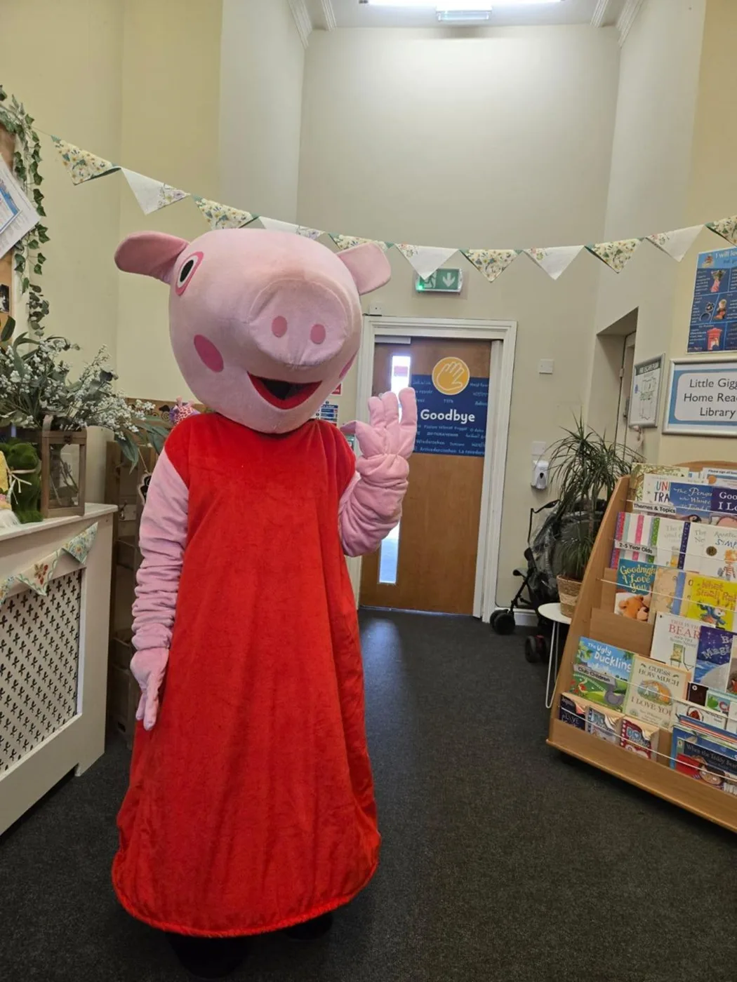 A Peppa Pig costume character in a red dress waving in the nursery hallway under bunting