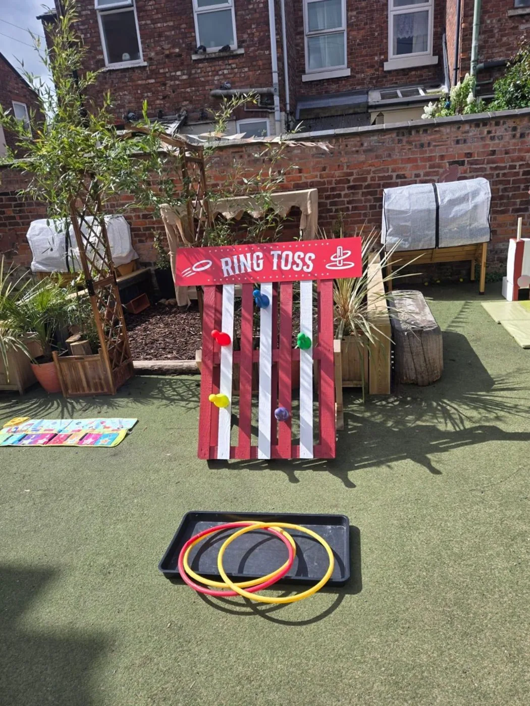A red and white striped ring toss board with pegs, with a tray of coloured hoops in front, in the nursery garden