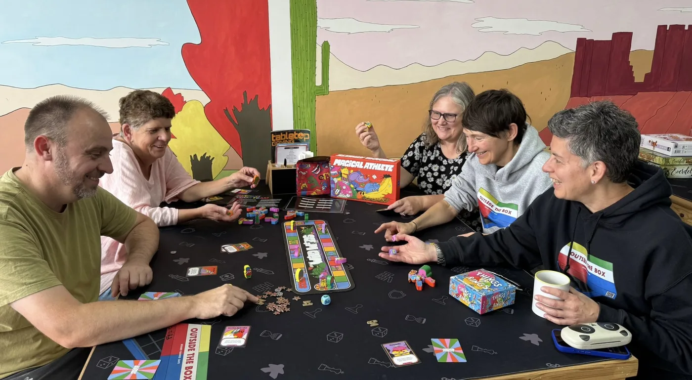 Five people around a table playing a colourful board game in front of a painted wall mural